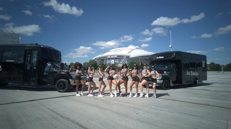 Dance team of nine women in matching black-and-white striped crop tops and shorts posing between two black shuttle buses in a sunny stadium parking lot with a large domed stadium and blue sky in the background.