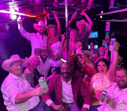 Group of adults in cowboy hats and party outfits cheering with drinks inside a neon-pink party bus around a center pole