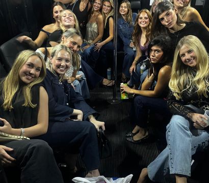 Group of smiling young women on a nighttime party bus interior, seated around a center pole and holding drinks during a night out.