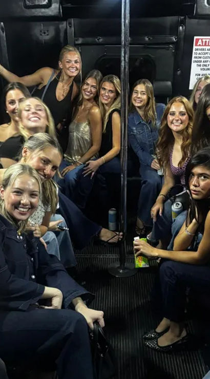 Cheerful group of women inside a party bus gathered around a center pole, holding drinks and enjoying a girls' night out nightlife ride.