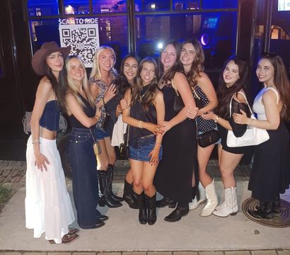 Ten friends in evening outfits and cowboy boots posing arm‑in‑arm in front of a blue‑lit party bus on a brick downtown sidewalk at night.