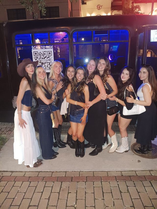 Ten friends in evening outfits and cowboy boots posing arm‑in‑arm in front of a blue‑lit party bus on a brick downtown sidewalk at night.