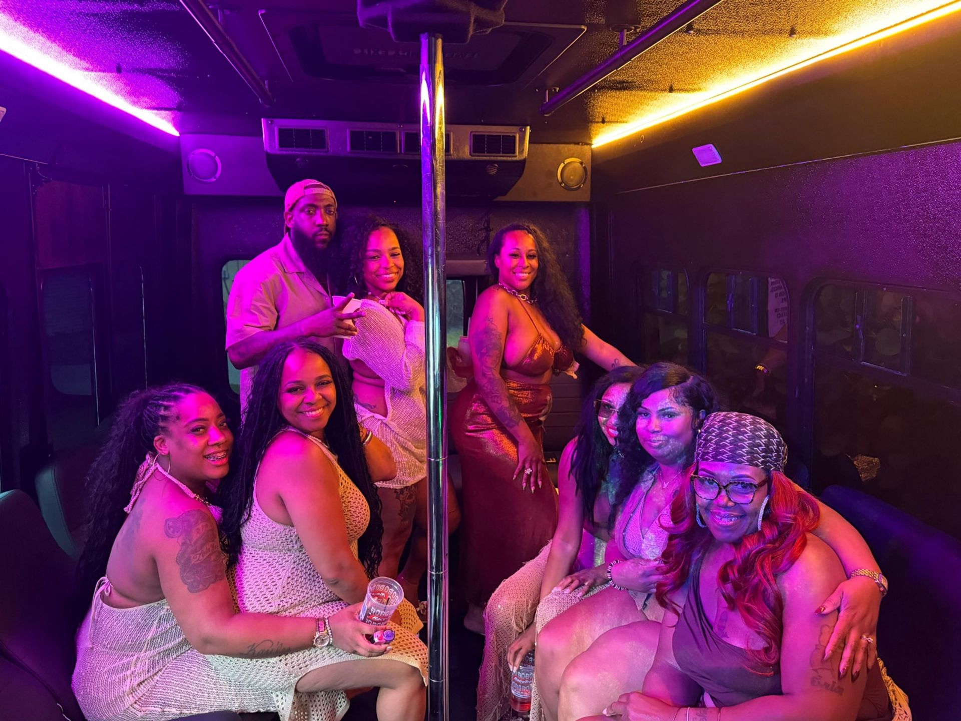Smiling group of friends inside a neon-lit party bus gathered around a chrome dance pole, wearing colorful party outfits and holding drinks.