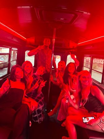 Group of young adults smiling and pointing inside a red-lit party bus with a central dance pole and city street visible through the windows