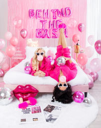Playful pink party scene: two people in matching hot-pink outfits lounging on a blush bed—one upside-down—both wearing white heart-shaped sunglasses and blowing bubblegum, surrounded by pink balloons, disco balls, a sequin lip pillow, foil fringe backdrop and metallic balloon letters for a celebratory vibe.