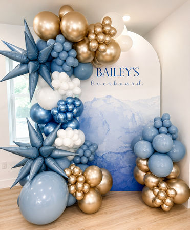 Indoor nautical-themed photo backdrop with cascading balloon arch in slate blue, sky blue, white and metallic gold, spiky star balloons and clustered round accents — stylish event decor.