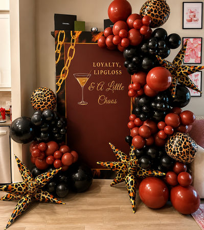 Cocktail-themed party backdrop reading 'Loyalty, Lipgloss & A Little Chaos' framed by black and deep red balloon garlands and leopard-print star inflatables on a wood floor