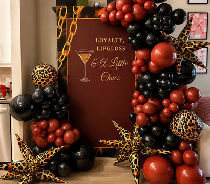 Cocktail-themed party backdrop reading 'Loyalty, Lipgloss & A Little Chaos' framed by black and deep red balloon garlands and leopard-print star inflatables on a wood floor