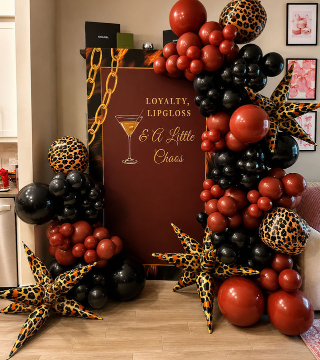 Cocktail-themed party backdrop reading 'Loyalty, Lipgloss & A Little Chaos' framed by black and deep red balloon garlands and leopard-print star inflatables on a wood floor