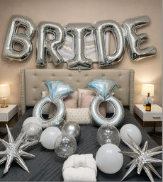 Silver foil "BRIDE" balloons over a tufted bed with giant diamond-ring balloons, silver and white balloons, star-shaped foil accents and a champagne bottle — bridal bedroom party decor.