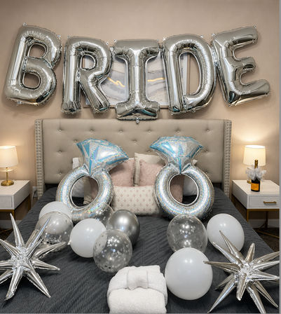 Silver foil "BRIDE" balloons over a tufted bed with giant diamond-ring balloons, silver and white balloons, star-shaped foil accents and a champagne bottle — bridal bedroom party decor.