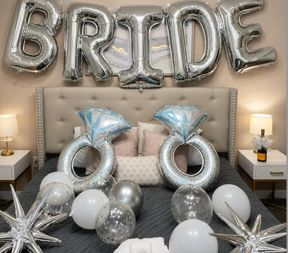 Silver foil "BRIDE" balloons over a tufted bed with giant diamond-ring balloons, silver and white balloons, star-shaped foil accents and a champagne bottle — bridal bedroom party decor.