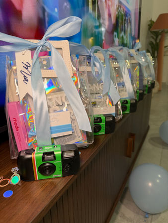 Row of clear gift bags tied with pastel blue ribbons containing green disposable cameras and holographic party favors on a wooden console, with light-blue balloons nearby