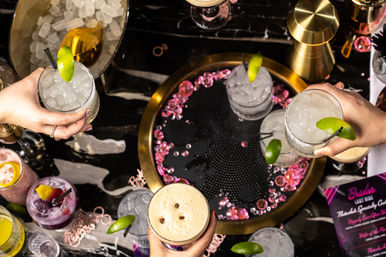 Top-down shot of a lively bar scene with hands holding lime-garnished crushed-ice cocktails, a frothy espresso drink, pink decorative gems and a gold tray on a black marble countertop.