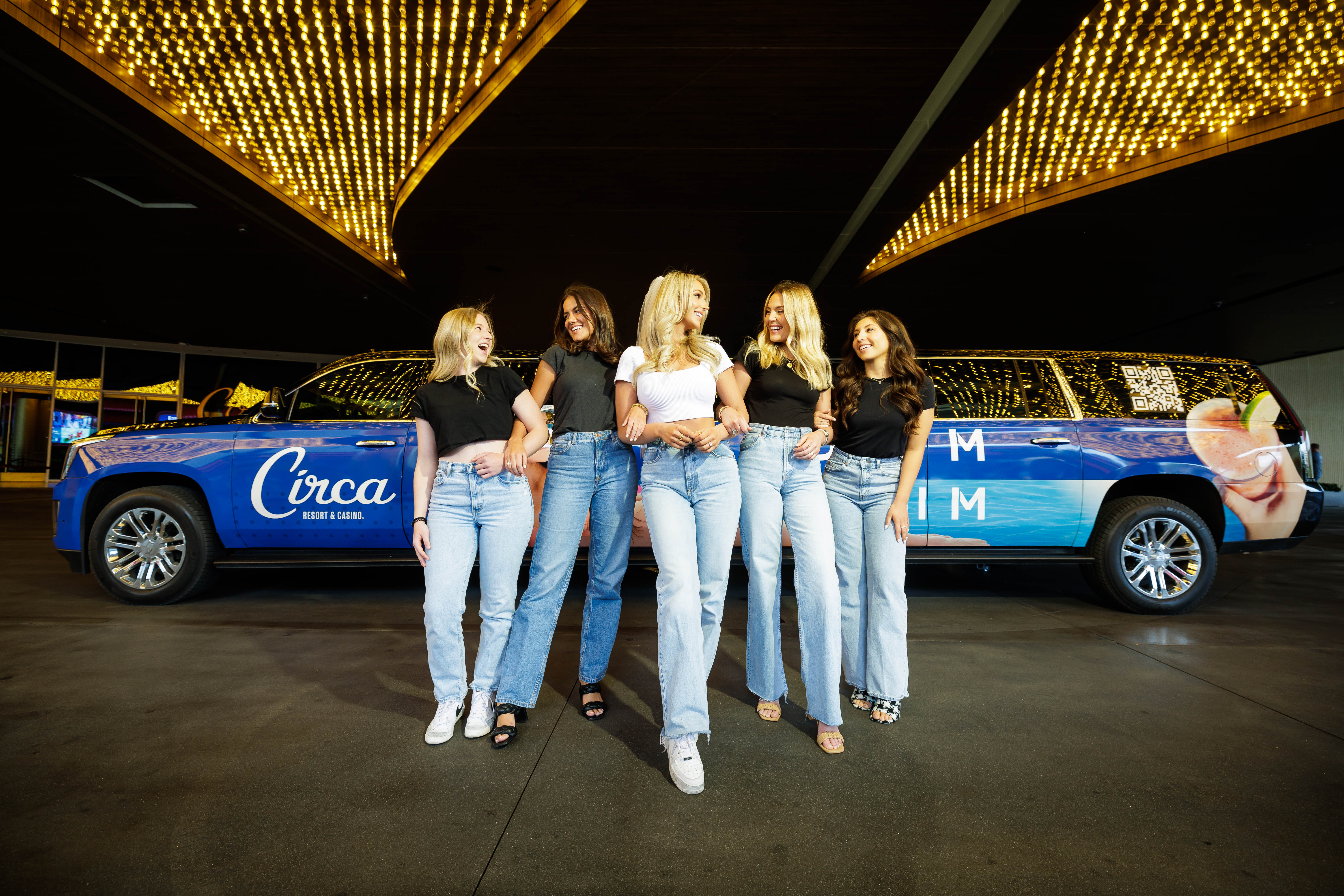 Five friends in casual black and white tops and light-wash jeans arm-in-arm, laughing as they walk past a wrapped blue stretch SUV beneath bright marquee lights at a casino entrance