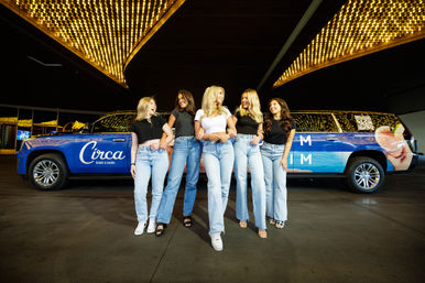 Five friends in casual black and white tops and light-wash jeans arm-in-arm, laughing as they walk past a wrapped blue stretch SUV beneath bright marquee lights at a casino entrance