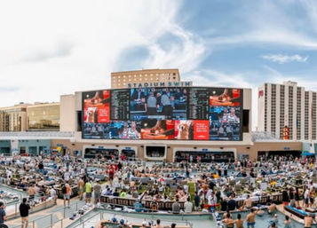 Crowded outdoor resort pool and lounge area with a giant stadium-style LED video wall showing sports, people in swimwear on lounge chairs and in pools, and high-rise hotels in the background under a blue sky.