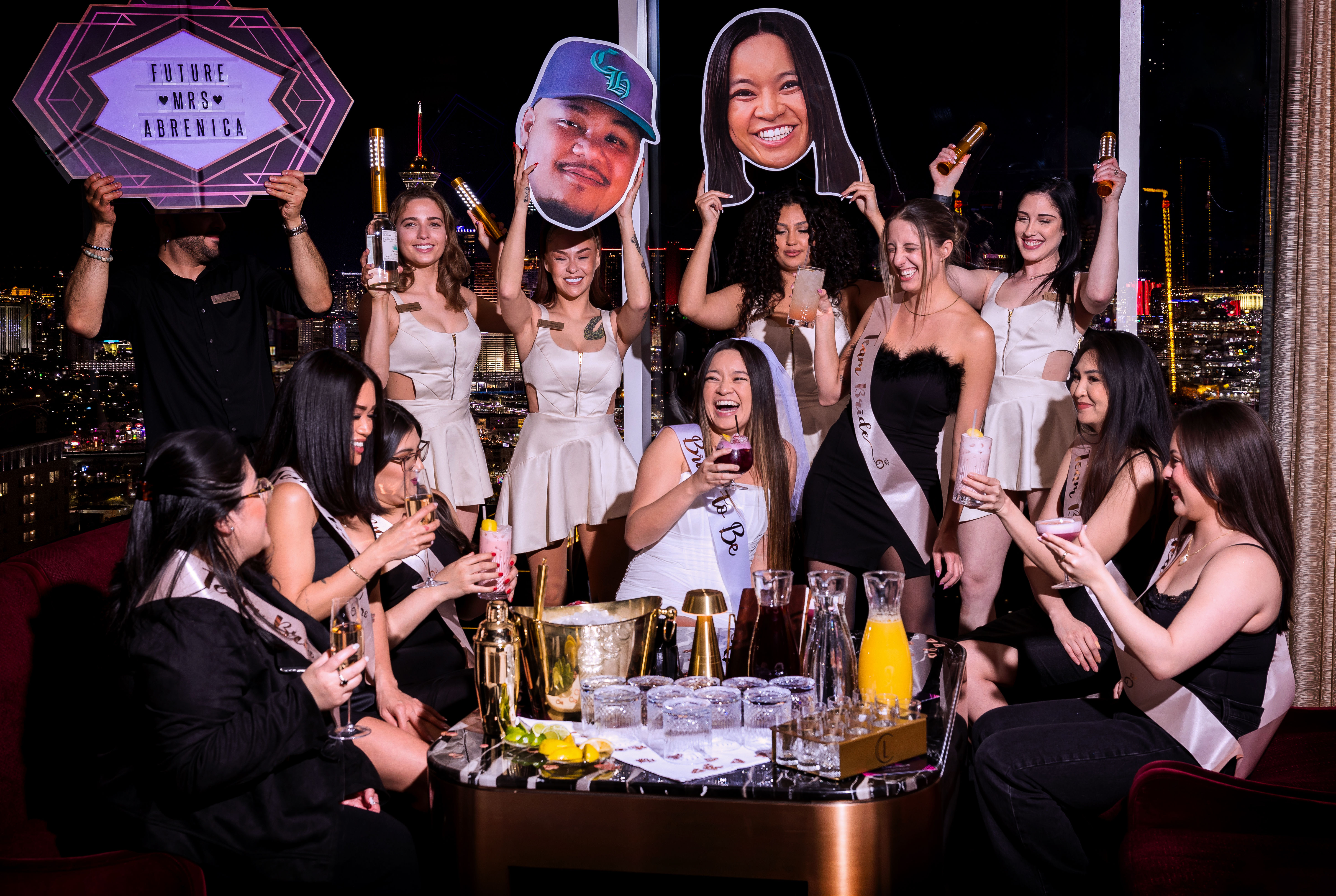 Bachelorette party at a rooftop lounge — bride-to-be in a white sash and veil laughing with friends in sashes, toasting cocktails around a table with champagne, pitchers and oversized face cutouts against a nighttime city skyline.
