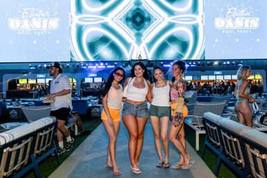 Four women smiling and posing on an outdoor pool deck at a lively nighttime pool party, with a DJ booth, large colorful LED screen, lounge chairs and illuminated bar area in the background — summer nightlife pool event.