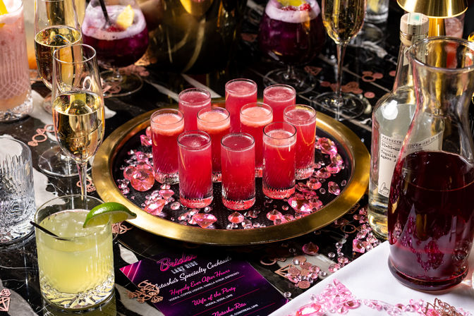 Vibrant party table with a gold tray of bright pink cocktail shots, champagne flutes, a lime margarita, a carafe of red punch and scattered pink gems and a party menu card.