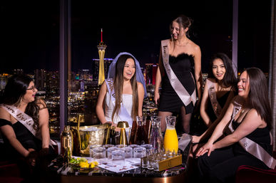 Bachelorette party: five women in sashes, a veiled bride laughing around a table with champagne bucket, glasses and pitchers, against a nighttime Las Vegas skyline with the Stratosphere Tower visible.