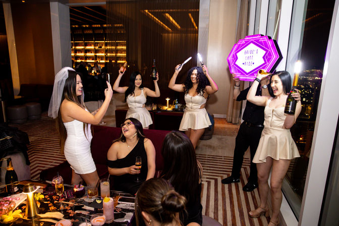 Bachelorette party in a stylish hotel suite at night — bride in a white veil celebrating with friends in matching cream dresses, popping champagne and waving sparklers beside a neon "Bride's Last Ride" sign with city lights visible through floor-to-ceiling windows