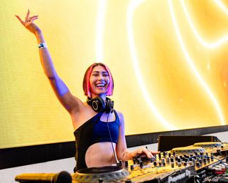 Smiling DJ with pink-streaked bob and headphones raising an arm while mixing on a professional DJ controller against a vibrant yellow LED stage backdrop.