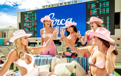 Five women in colorful swimsuits and pink cowboy hats cheering with champagne on a sunny stadium pool deck with lounge seating and a large LED screen in the background