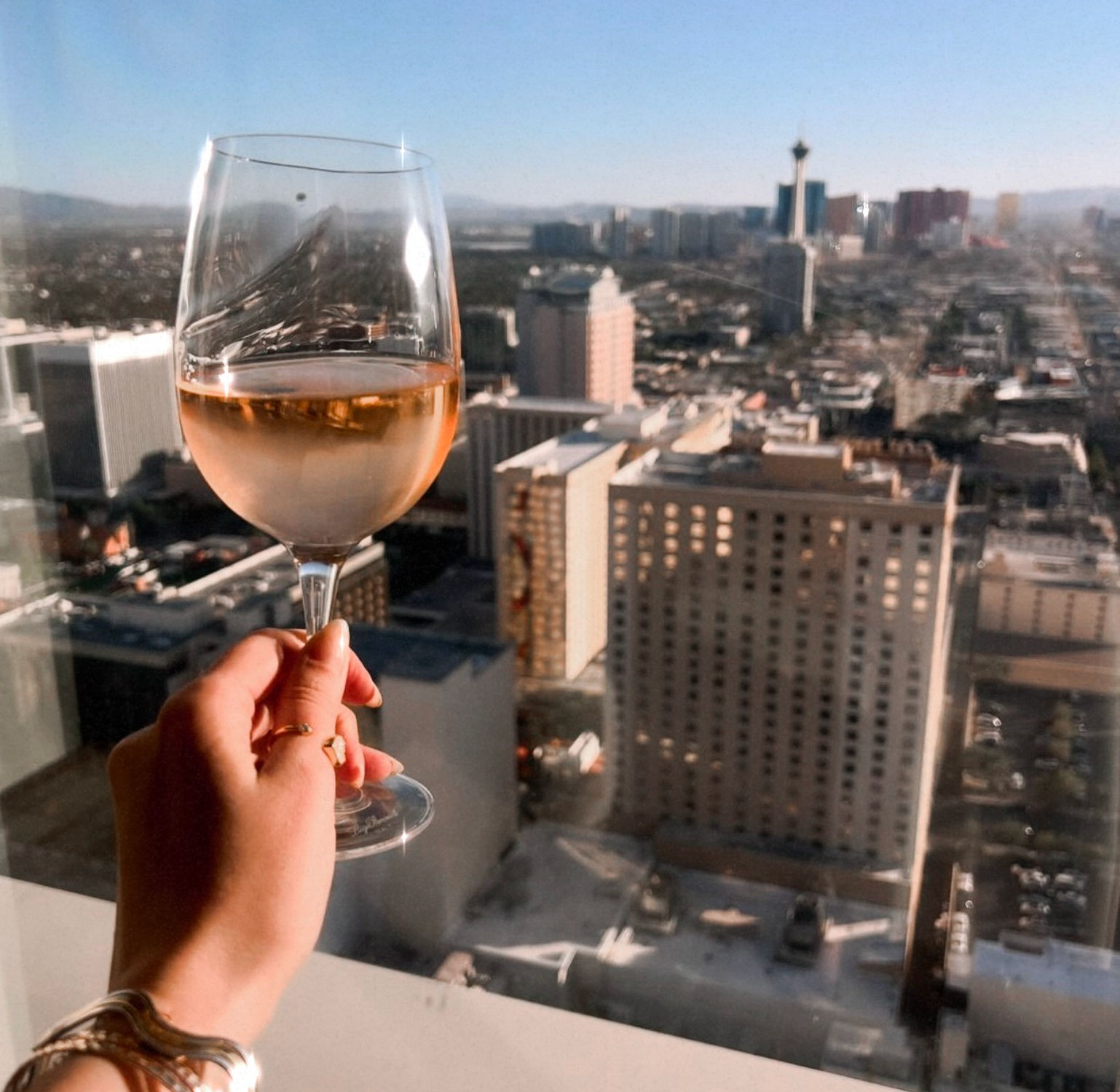 Hand holding a rosé wine glass against a sunlit high-rise city skyline with a distant observation tower — relaxed rooftop view.