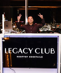 Smiling DJ at a rooftop cocktail bar booth with a laptop, flashing peace signs against a sparkling city skyline at night