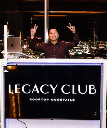 Smiling DJ at a rooftop cocktail bar booth with a laptop, flashing peace signs against a sparkling city skyline at night