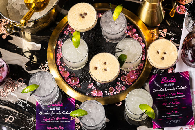 Overhead shot of party-ready cocktails on a gold tray at a bachelorette celebration — espresso martinis with coffee bean garnish, crushed-ice lime drinks, pink gem decor and event menus on a black marble table.