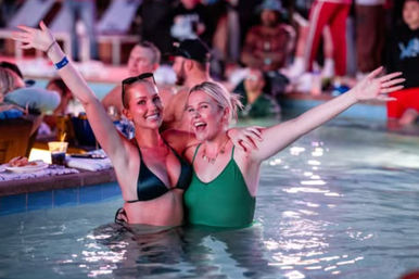 Two friends in swimsuits smiling and cheering with arms raised while standing in a brightly lit pool at a lively nighttime pool party, guests and drinks visible in the background.