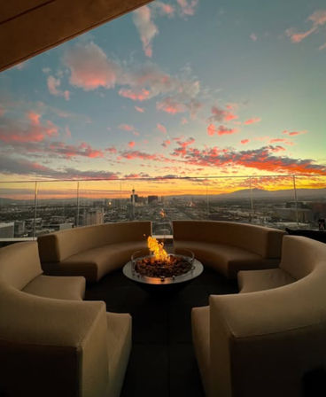 Cozy rooftop lounge with curved beige sofas encircling a glowing fire pit, glass railing framing an urban skyline beneath a pink-orange sunset over distant hills