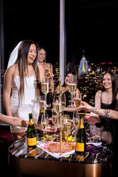 Bride-to-be in a white mini dress and veil laughing with girlfriends during a rooftop bachelorette toast around a tiered champagne tower, bottles and city skyline lights glowing at night.