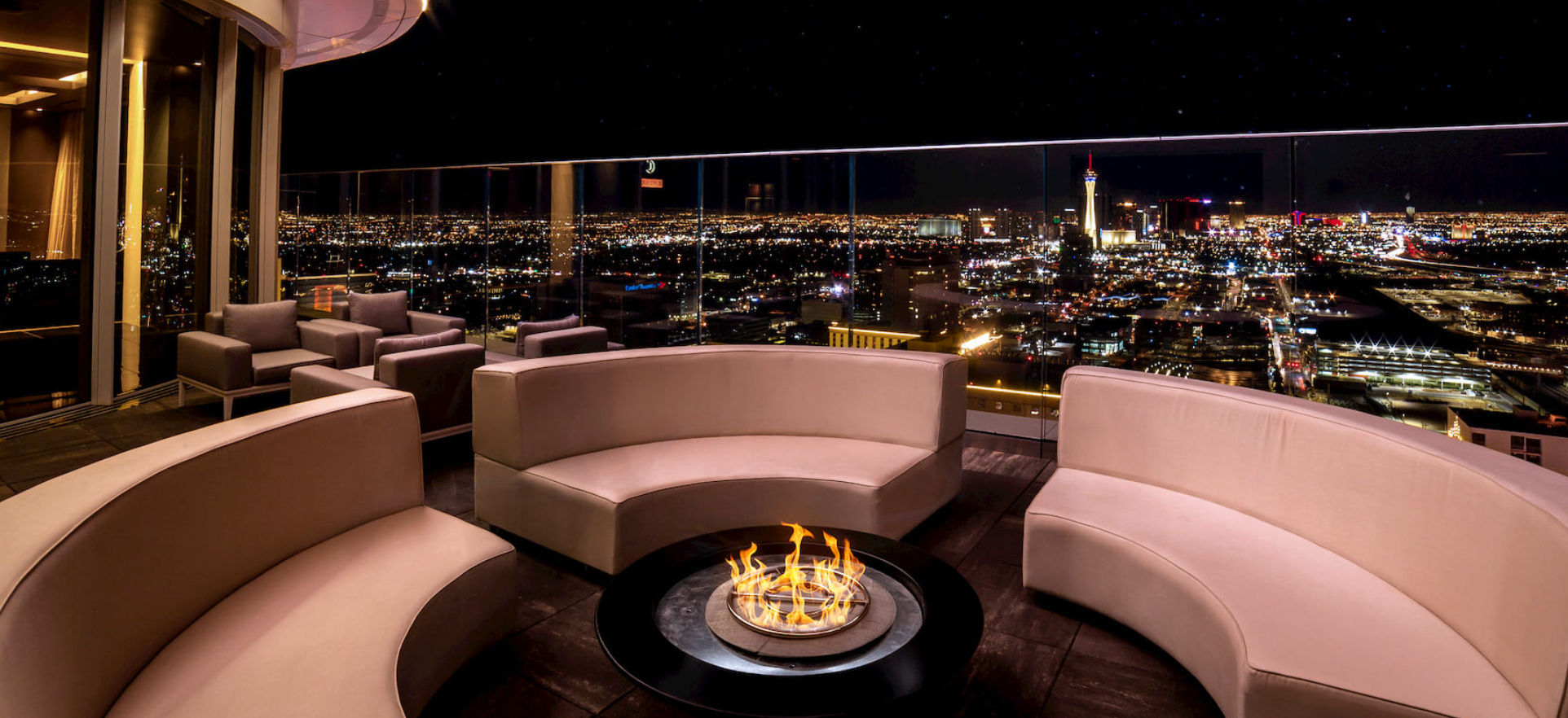 Chic rooftop lounge with curved beige sofas around a central fire pit, glass railing overlooking the brightly lit Las Vegas skyline at night