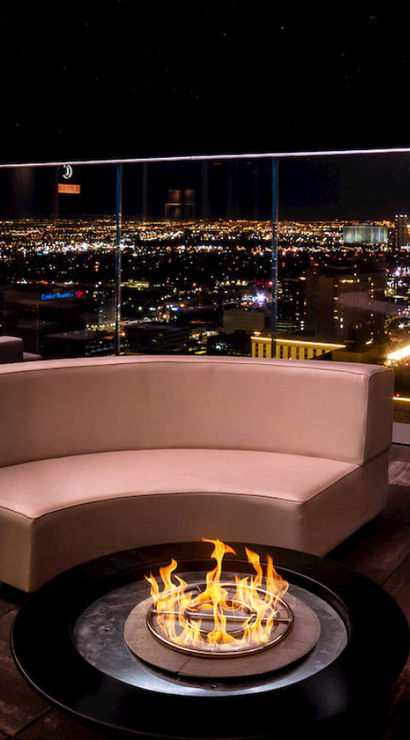 Chic rooftop lounge with curved beige sofas around a central fire pit, glass railing overlooking the brightly lit Las Vegas skyline at night