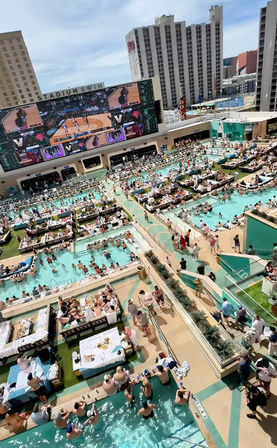 Sunlit rooftop resort pool deck with multiple pools, cabanas and daybeds, a huge outdoor video screen showing basketball, and crowds of sunbathers amid high-rise hotels.