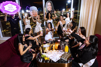 Bachelorette party in a high-rise hotel suite, bride in a white sash laughing with friends toasting cocktails and holding oversized face cutouts against a glittering city skyline at night.