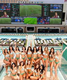 Group of women in matching green bikinis posing on a rooftop pool deck with rows of lounge cabanas and a giant multi-screen video wall showing sports