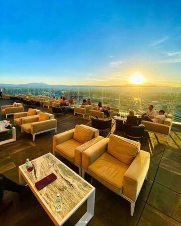 Sunset rooftop lounge with cozy tan armchairs and marble-top tables on a glass-railed terrace, people socializing with panoramic city skyline and glowing horizon view.