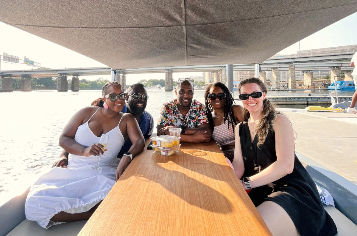 Five friends smiling at a wooden table on a sunny boat ride under a canopy, enjoying drinks and fruit cups with a river, bridge and urban waterfront in the background