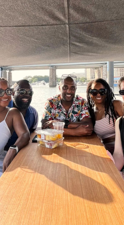 Five friends smiling at a wooden table on a sunny boat ride under a canopy, enjoying drinks and fruit cups with a river, bridge and urban waterfront in the background