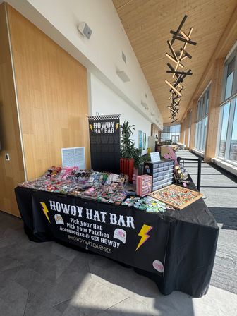 Colorful hat patches, pins and accessories on a vendor table at a sunlit upper-floor pop-up market with floor-to-ceiling windows, city skyline views and warm wooden ceiling.