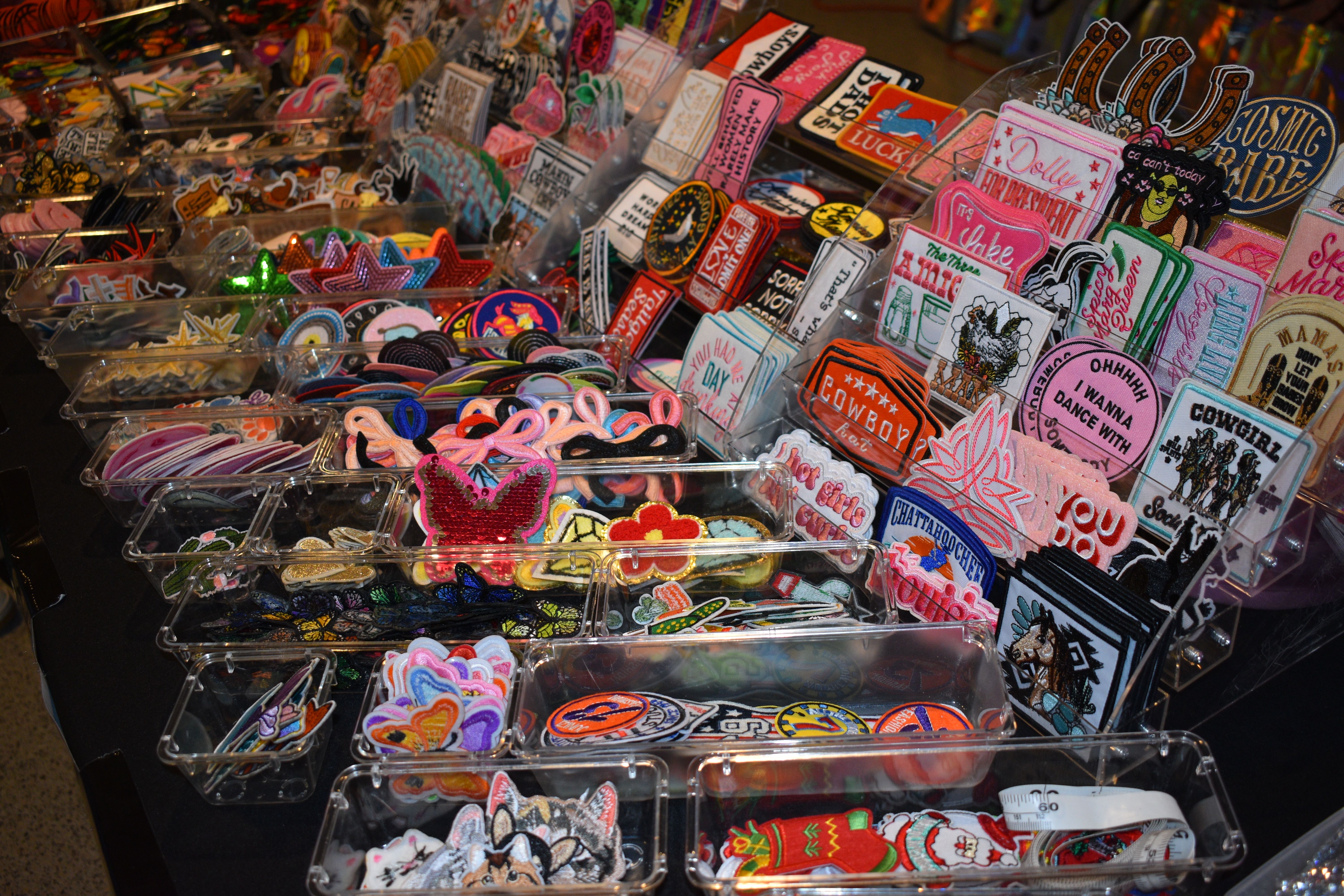 Vibrant embroidered iron-on patches and appliqués in clear bins and tiered racks on a craft market table, showing hearts, stars, animals and playful text designs.