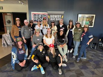 Group of smiling women in a stadium lounge wearing colorful baseball caps and team jerseys, posing together in front of sports-themed signage.