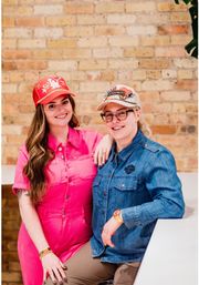 Two smiling women wearing decorated baseball caps pose against a light urban brick wall — one in a bright pink jumpsuit, the other in a denim shirt and glasses, casual indoor portrait.