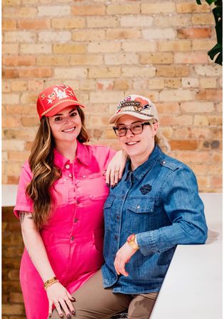 Two smiling women wearing decorated baseball caps pose against a light urban brick wall — one in a bright pink jumpsuit, the other in a denim shirt and glasses, casual indoor portrait.