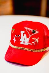 Red trucker hat decorated with a white 'LA' patch, martini glass patch with olive, gold airplane pin, heart-with-eyes and star patches, tiny cherries pin and a gold chain trim across the brim — stylish Los Angeles-themed accessory.