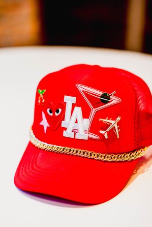 Red trucker hat decorated with a white 'LA' patch, martini glass patch with olive, gold airplane pin, heart-with-eyes and star patches, tiny cherries pin and a gold chain trim across the brim — stylish Los Angeles-themed accessory.